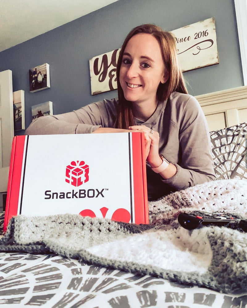 Young adult woman sitting in bed holding a SnackBOX and smiling.